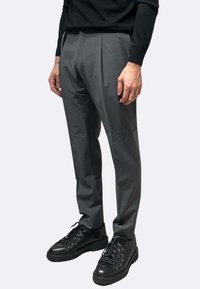 Pantaloni grigi su misura con pieghe, realizzati in un tessuto liscio. Abbinati a scarpe nere robuste con lacci e superficie testurizzata.