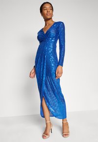 Μπλε φόρεμα με μακριά μανίκια, βαθύ V-ντεκολτέ, καλυμμένο με παγιέτες, με πλαϊνό σκίσιμο και ρούχα που πέφτουν ωραία, συνδυασμένο με γυμνές πέδιλες ψηλές.