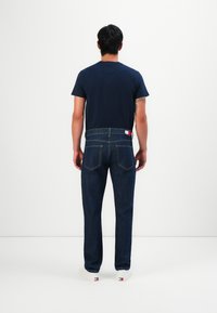 Homme de dos portant un t-shirt bleu marine, un pantalon en jean bleu foncé avec des poches arrière et des baskets blanches avec des détails de marque au talon.