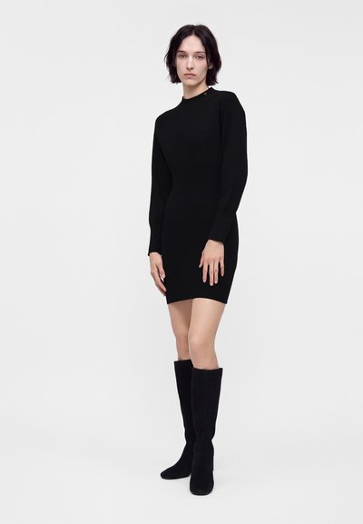 Femme debout portant une robe mini noire ajustée à manches longues et des bottes noires montantes sur fond clair uni.