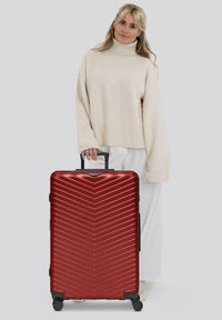 Mujer con un jersey de cuello alto beige sosteniendo el asa de una gran maleta rígida roja con estrías diagonales, de pie sobre un fondo gris.