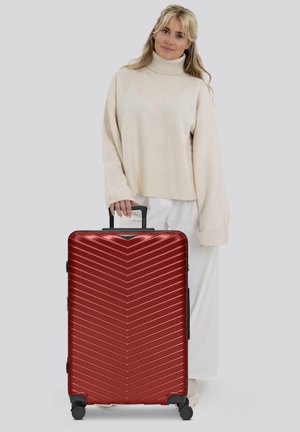 Mujer con un jersey de cuello alto beige sosteniendo el asa de una gran maleta rígida roja con estrías diagonales, de pie sobre un fondo gris.