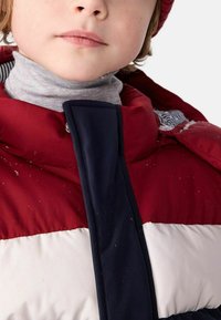 Doudoune rouge et marine avec une bande blanche, col haut et fermeture à pression en argent. Portée sur un col roulé gris, elle présente une texture douce.