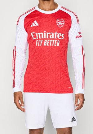 Uomo che indossa la maglia da calcio rossa e bianca dell'Arsenal con il logo Emirates Fly Better e pantaloncini bianchi Adidas.