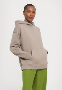 Haut à capuche beige surdimensionné avec une poche frontale et un cordon de serrage, associé à un pantalon vert avec une texture lisse.