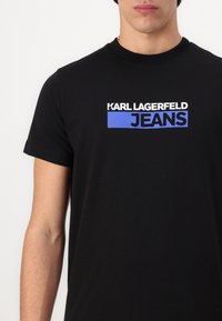 Juodas medvilninis marškinėlis trumpomis rankovėmis, su baltu ir mėlynu „KARL LAGERFELD JEANS“ logotipu krūtinėje. Lygus paviršius.