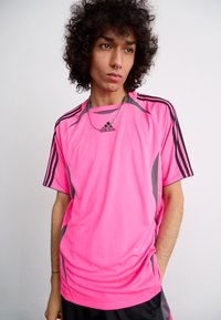 Ryškiai rožinė sportinė marškinė su juodais akcentais, trumpomis rankovėmis ir Adidas logotipu. Marškiniai turi tekstūruotą paviršių ir kontrastingas šonines juostas.