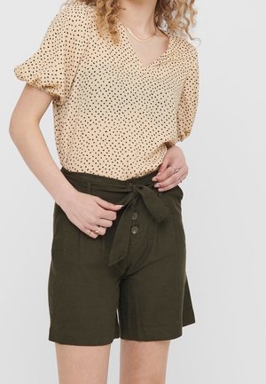 Femme portant un chemisier beige à manches courtes avec des pois noirs et un short vert foncé taille haute noué avec une ceinture en tissu.