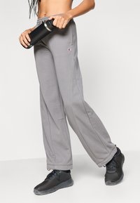 Γκρι φούτερ παντελόνια με άνετη εφαρμογή και ορατές ραφές, σε συνδυασμό με μαύρα αθλητικά παπούτσια με δίχτυ και μαύρο μονωμένο παγούρι.