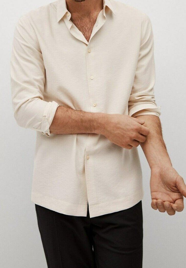 Homme retroussant les manches d'une chemise boutonnée couleur crème, portant un pantalon noir, sur un fond clair uni.