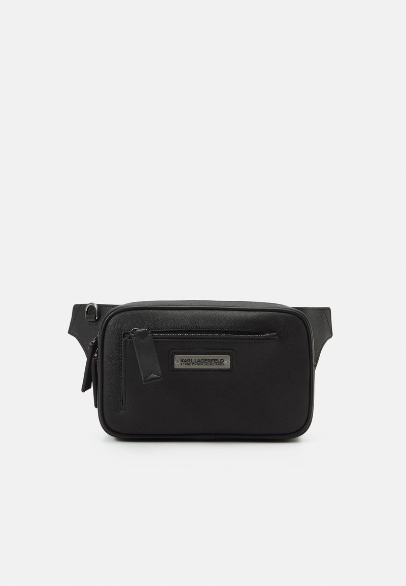 Sac en tissu noir à bandoulière avec une texture lisse, des compartiments zippés et une plaque de logo argentée. Il est doté d'une sangle réglable détachable.