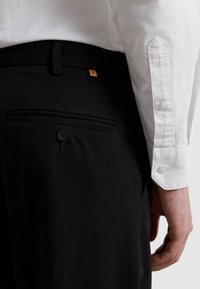 Czarne spodnie o gładkiej fakturze, z tylną kieszenią zapinaną na guzik i pomarańczową metką z logo. W połączeniu z białą koszulą zapinaną na guziki.