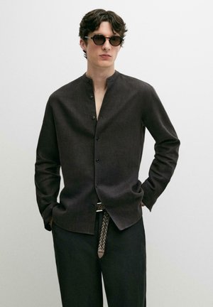 Młody mężczyzna w okrągłych okularach przeciwsłonecznych, ciemnej koszuli na guziki z kołnierzem typu band, plecionym paskiem i ciemnych spodniach, stojący z rękami w kieszeniach.