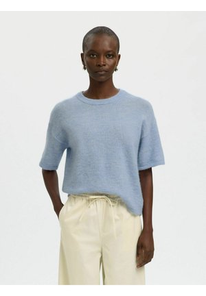 Žena tamne puti s kratkom kosom koja nosi svijetloplavi kratki pulover s pufastim izgledom i kremaste hlače sa vezicama, na jednobojnoj pozadini.