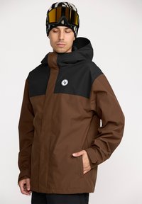 Brun og sort vandtæt jakke med hætte, der har et logo foran, sidelommer og justerbare manchetter, lavet af slidstærkt stof.