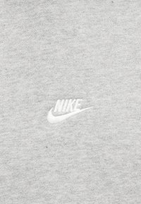 Λευκό λογότυπο Nike κεντημένο σε ανοιχτογκρι υφασμάτινο φόντο, εμφανίζοντας καθαρά το όνομα της μάρκας και το σύμβολο swoosh.