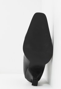 Chaussure à talon haut en cuir noir avec un bout carré reposant côté semelle contre un mur d’angle blanc et un sol.