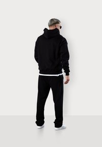 Schwarzer Hoodie mit hohem Kragen und gerippten Bündchen, kombiniert mit schwarzen Jogginghosen. Das Modell trägt weiße Sneaker. Der Hintergrund ist schlicht.