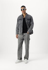 Chaqueta de mezclilla gris con dos bolsillos en el pecho, combinada con pantalones de mezclilla gris a juego. Zapatillas blancas y gafas de sol completan el conjunto.