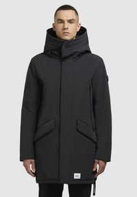 Parka neagră cu guler înalt, buzunare laterale cu fermoar, buzunare mari în față, material neted și etichetă de brand subtilă la tiv.