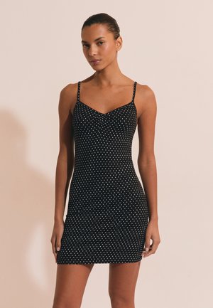 Kobieta ubrana w czarną mini sukienkę bez rękawów w białe groszki, stojąca na jednolitym beżowym tle.