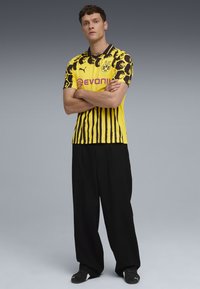 Cămașă de fotbal cu dungi galbene și negre, având un model îndrăzneț cu imprimeu de leopard. Prezintă mâneci scurte și accente cu logo. Asortată cu pantaloni largi negri.