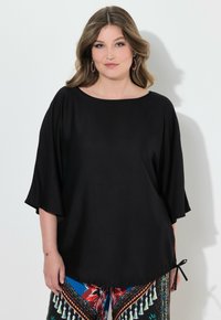 Top nero dalla vestibilità ampia con scollatura larga e maniche a tre quarti; presenta un dettaglio con fiocco all'orlo. Tessuto morbido e liscio.