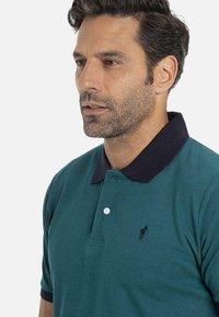 Homme avec des cheveux courts et foncés et une barbe portant un polo teal avec un col noir et un petit logo brodé sur la poitrine, sur fond blanc.