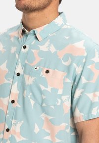 Camicia a maniche corte con bottoni di colore azzurro chiaro, decorata con motivi astratti in rosa e bianco. Presenta una tasca sul petto e bottoni marroni sul davanti.
