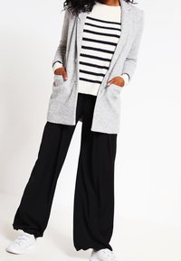 Cárdigan largo gris con bolsillos sobre un suéter a rayas crema y azul marino, combinado con pantalones negros holgados y zapatillas blancas.