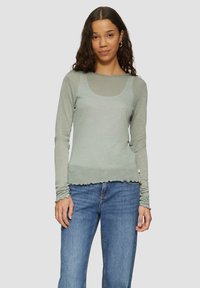 Hellgrünes Langarmshirt mit Rundhalsausschnitt und gewelltem Saum, aus einem glitzernden, weichen Material. Kombiniert mit blauen Jeans.
