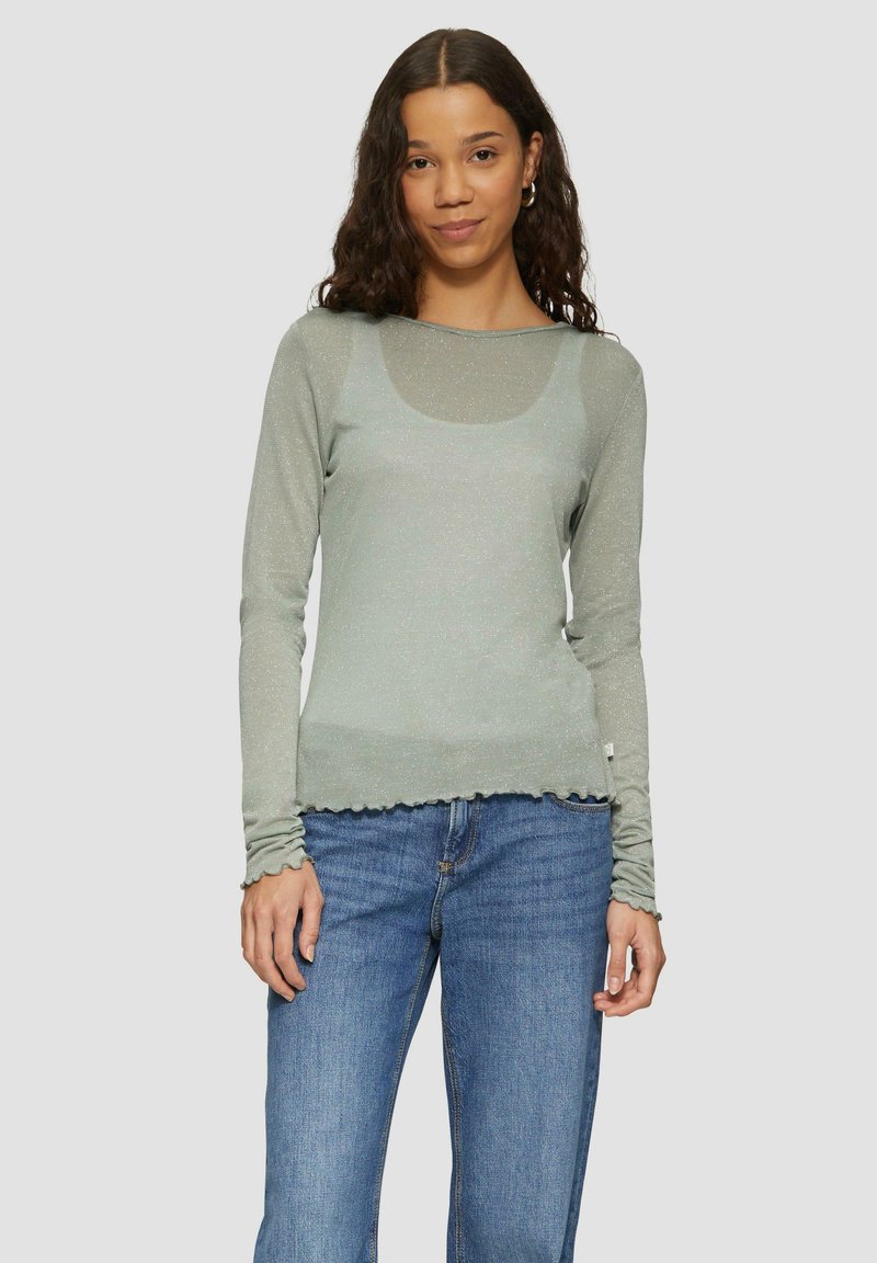 Hellgrünes Langarmshirt mit Rundhalsausschnitt und gewelltem Saum, aus einem glitzernden, weichen Material. Kombiniert mit blauen Jeans.