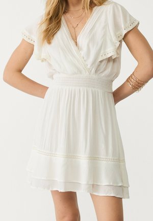 Vrouw draagt een witte korte mouwen jurk met V-hals en lagen, met een geplooide taille, kanten rand, gouden gelaagde kettingen en gouden armbanden om de rechter pols.