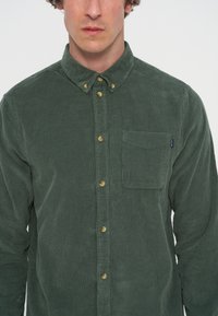 Homme portant une chemise en velours côtelé vert foncé avec une poche poitrine et un col boutonné, vu des épaules au menton.