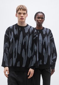 Pleteni puloverji v črni barvi z abstraktnim sivim vzorcem, z okroglim ovratnikom, dolgimi rokavi in rebrastimi manšetami, ki ju nosita dva modela.