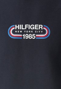 Testo "HILFIGER New York City 1985" con strisce curve rosse, bianche e blu su sfondo di tessuto scuro