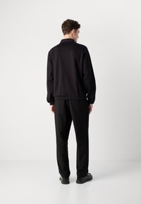 Schwarze Bomberjacke mit elastischem Saum und Bündchen, kombiniert mit locker sitzenden schwarzen Hosen, von hinten vor einem neutralen Hintergrund betrachtet.