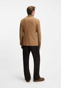 Blazer in lana marrone con uno spacco singolo e revers, abbinato a pantaloni neri e scarpe marroni, visto da dietro.