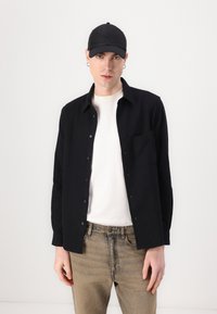 Black button-up shirt over a white t-shirt, paired with light brown patterned jeans and a black cap, featuring embroidered accents.