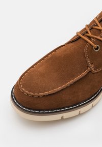 Zapato de ante marrón con puntera redonda, costuras en crema contrastantes y una suela de goma con patrones de tread visibles. Diseño de cordones y ojales.