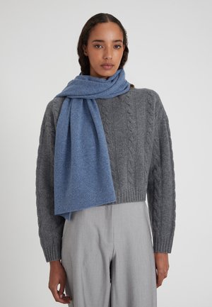 Une écharpe en laine bleue drapée sur un pull en mailles gris, assortie à un pantalon gris clair. L'écharpe présente une texture douce et un design simple.