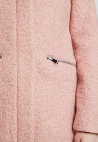 Primer plano de una persona vistiendo un abrigo texturizado rosa con un bolsillo de cremallera plateado visible en el lado derecho frontal.