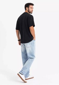 Černé tričko s krátkým rukávem, světle modré volné džíny, bílé tenisky s hnědými podrážkami, stojící pozice, zezadu s rukama v kapsách.