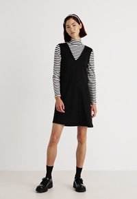 Vestido negro sin mangas con escote en V, llevado sobre una camiseta de manga larga a rayas. Presenta un corte casual y se combina con zapatos negros de cordones.