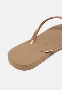Chinelo castanho com uma sola de borracha texturizada e tiras metálicas em cor dourado-rosa. A sola tem uma forma arredondada e uma ligeira elevação.
