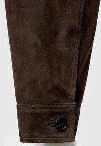Tessuto scamosciato marrone con una texture liscia. Presenta una chiusura a bottone e orlo cucito in modo ordinato sul polsino.