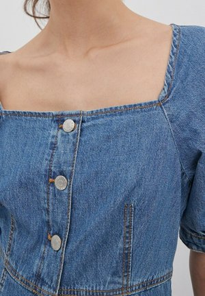 El vestido de denim presenta un escote cuadrado, mangas cortas abullonadas, botones plateados en el frente y costuras naranjas en contraste visibles.