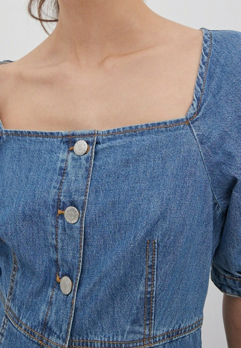Denim-mekko, jossa on neliönmuotoinen pääntie, lyhyet puffihihat, hopeiset napit edessä ja näkyvää oranssia ommelta.