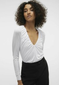 Maglietta bianca a maniche lunghe con scollo a V profondo e dettagli arricciati sul petto, abbinata a pantaloni neri sartoriali per un look a contrasto.