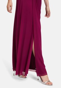 Dlhé magentové šaty s rozparkom po stehná; vyrobené z hladkej látky. Majú prúdny dizajn a sú skombinované s metalickými striebornými opätkami.
